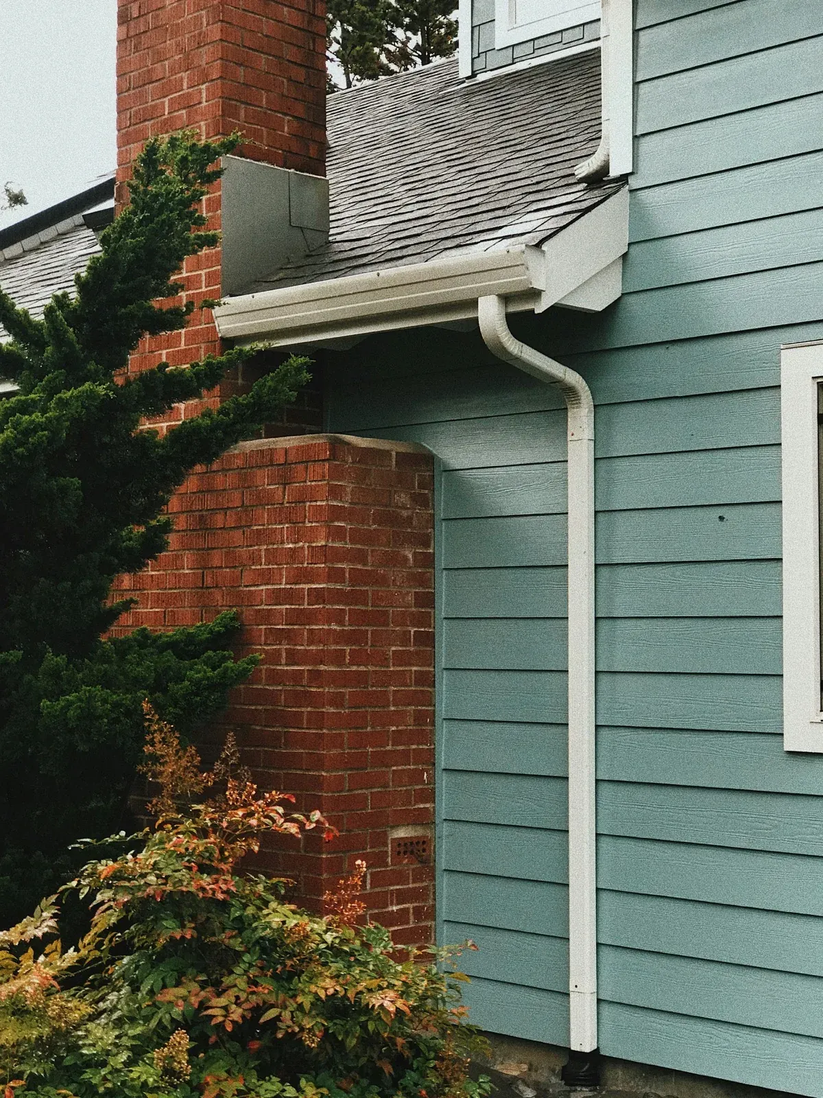 White seamless aluminum gutter and downspout installed on a blue-gray siding home with brick chimney in Lino Lakes, MN. Northern Forge Construction gutter installation and drainage solutions